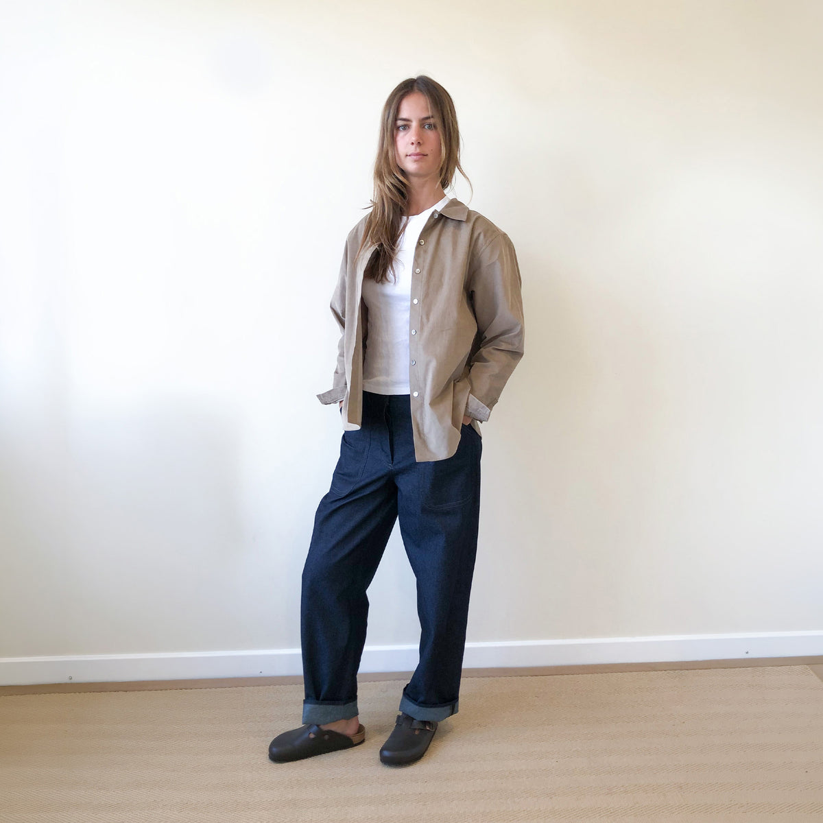 A woman stands in a minimalist room with a cream wall and light wood floor, wearing a beige overshirt over a white t-shirt, dark navy wide-leg trousers, and dark brown slip-on shoes.