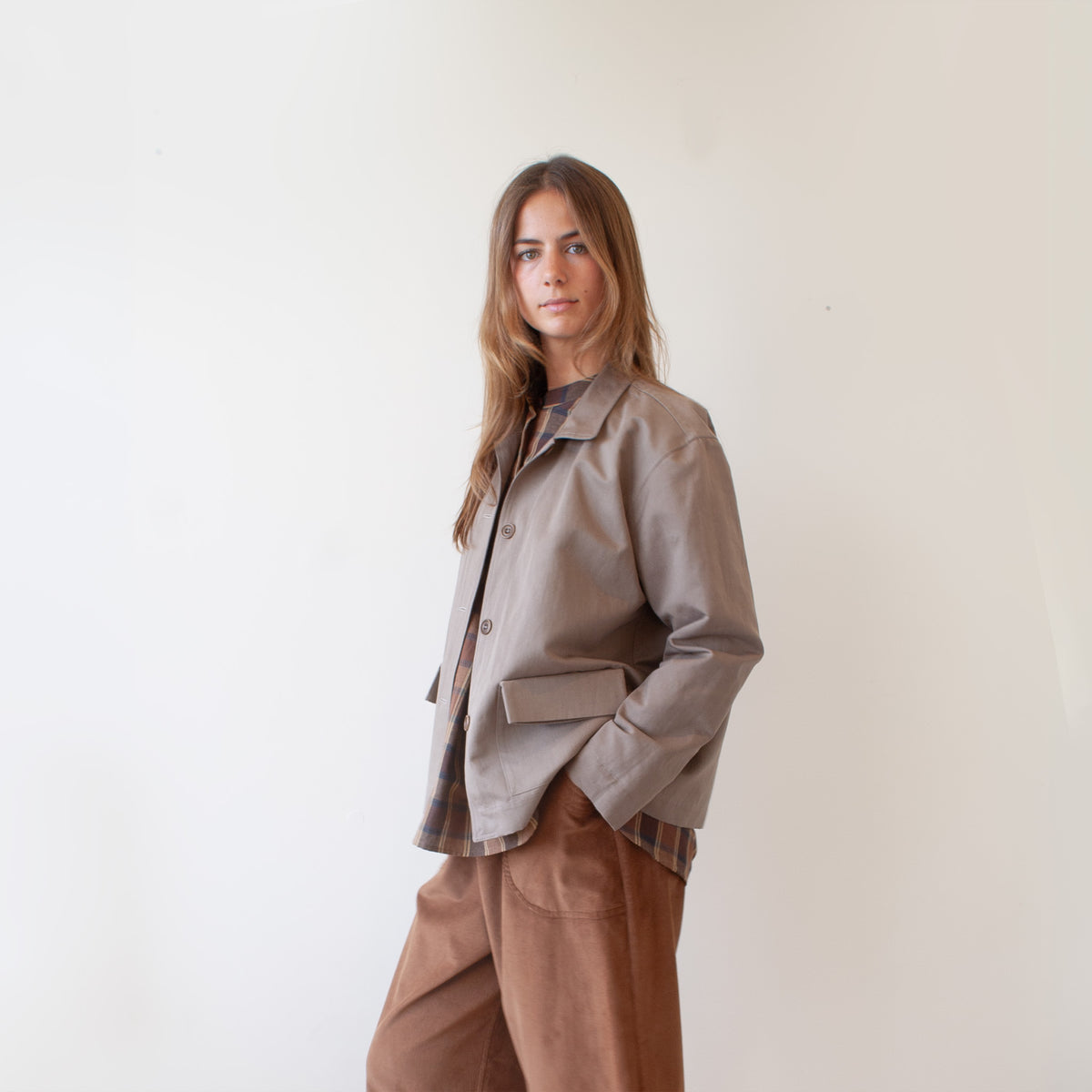 A woman modeling a light brown utility jacket over a plaid shirt and brown corduroy pants, standing against a plain off-white wall.