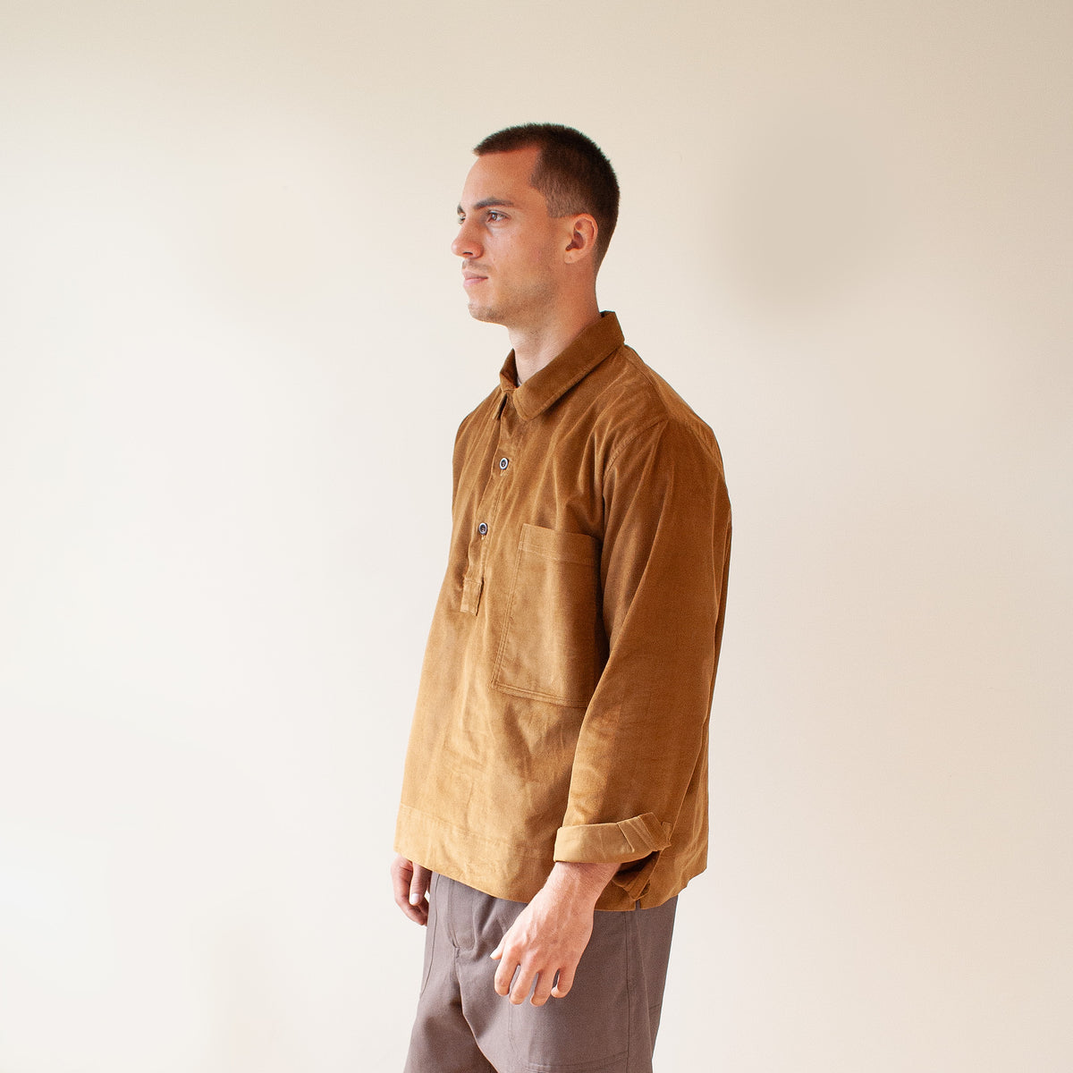 A man in profile wearing a brown corduroy button-up shirt and gray trousers, standing against a plain beige background.