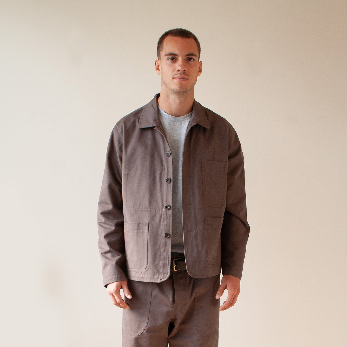 A man wearing a dark brown utility jacket over a light gray t-shirt and matching brown pants, standing against a plain beige background.