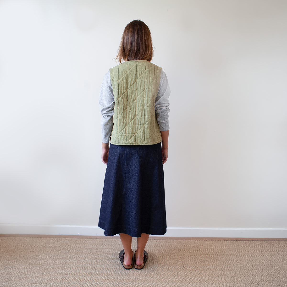 A woman stands with her back to the camera wearing a light sage green quilted vest over a white long-sleeve shirt and a dark navy midi skirt, in a minimalist room with a white wall and light wood floor.