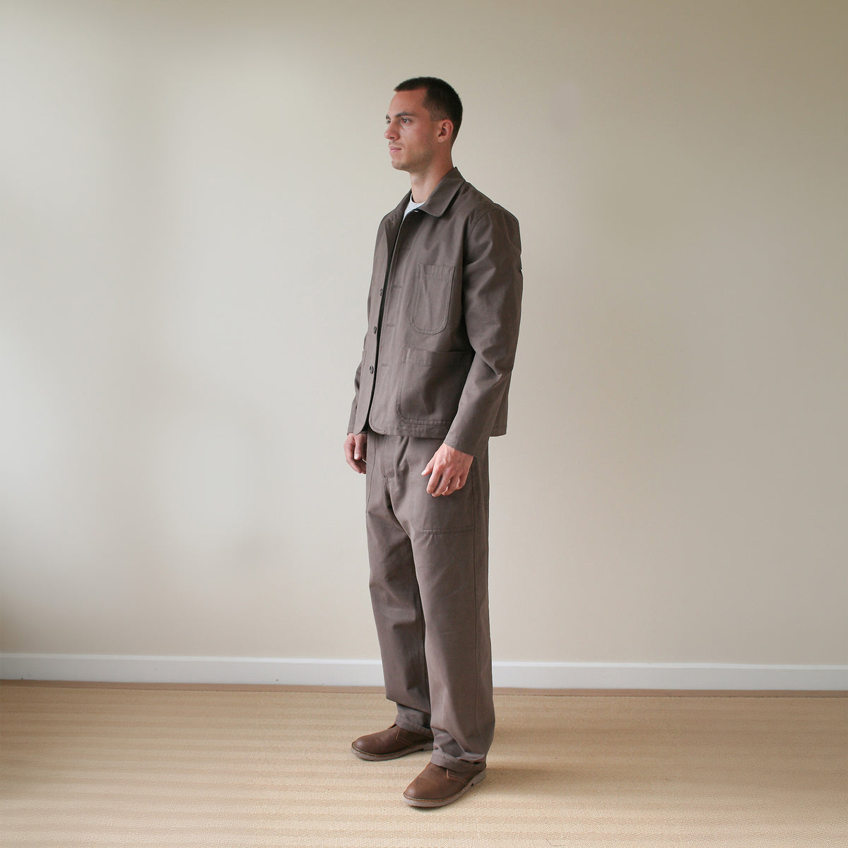 A man wearing a matching brown work jacket and trousers set, standing in profile against a plain beige wall and light wood floor.