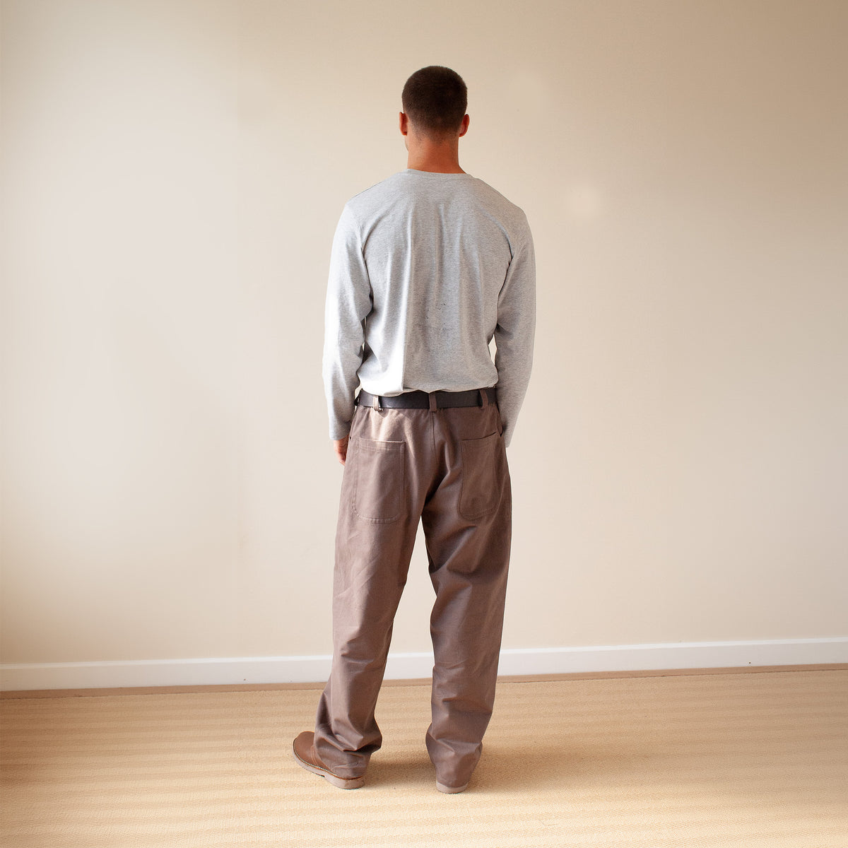 A man viewed from behind wearing a light gray long-sleeve shirt and brown pants, standing on a light wood floor against a plain beige wall.