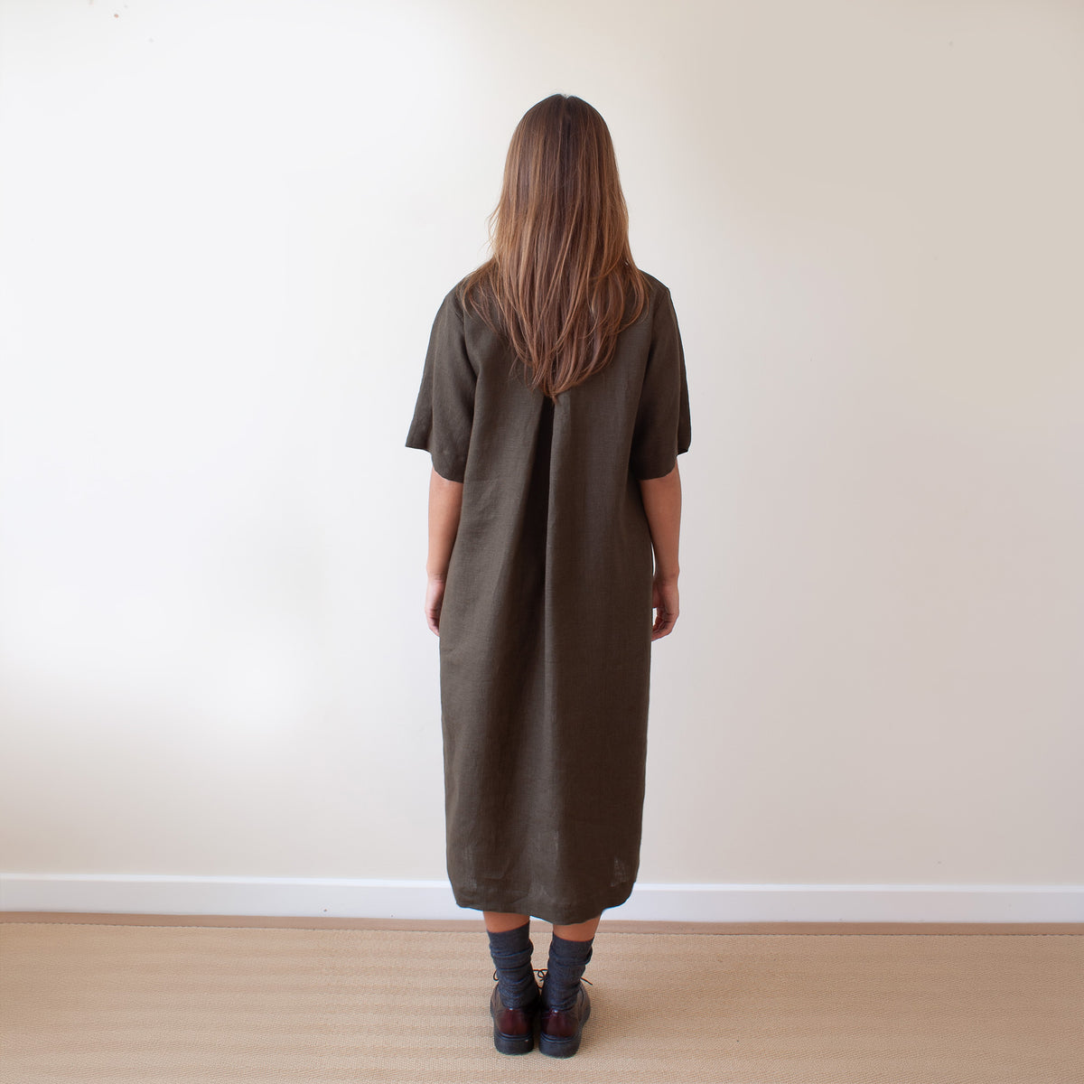 A woman viewed from behind wearing a loose, short-sleeved olive brown dress, standing on a light wood floor against a plain off-white wall.