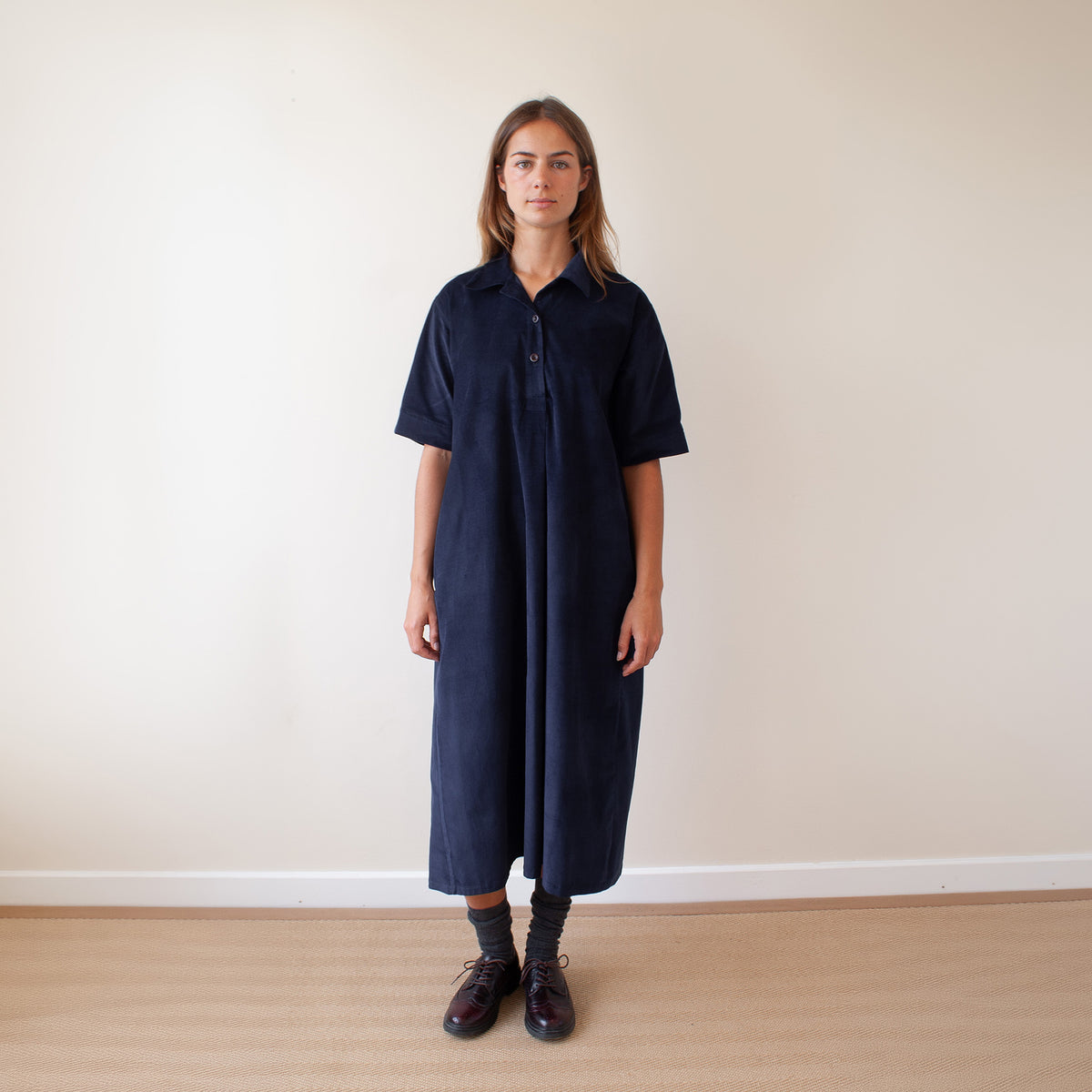 A woman stands in a room wearing a dark navy blue short-sleeve button-up dress, paired with dark socks and brown leather shoes, against a plain beige wall and light wood floor.