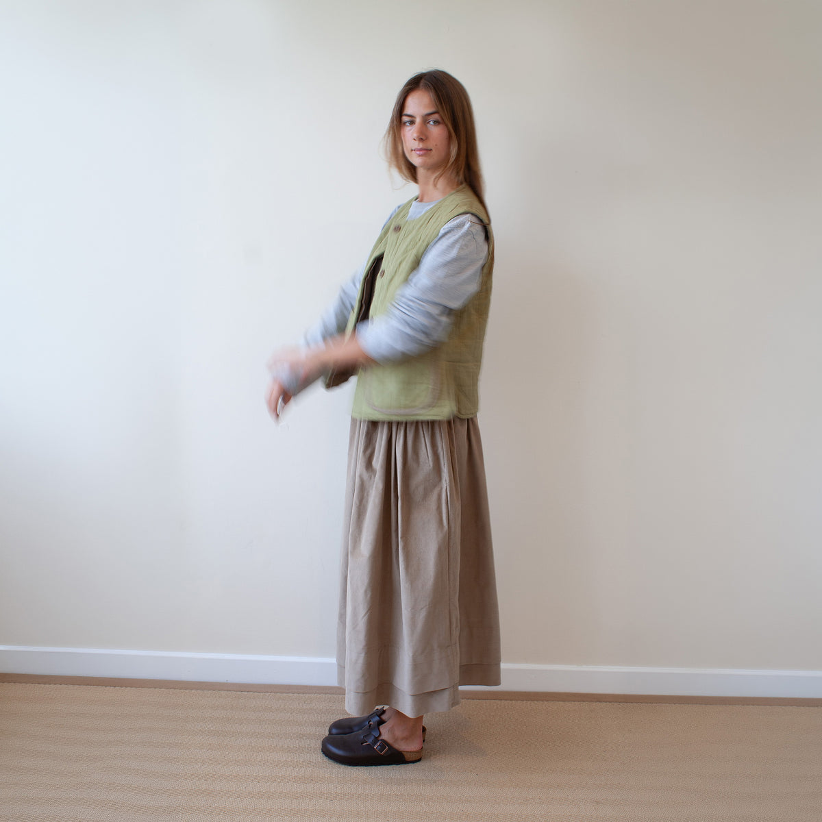 A woman stands in profile wearing a light olive green quilted vest over a light blue long-sleeve shirt, paired with a long taupe skirt and dark loafers, against a plain beige wall and light wood floor.