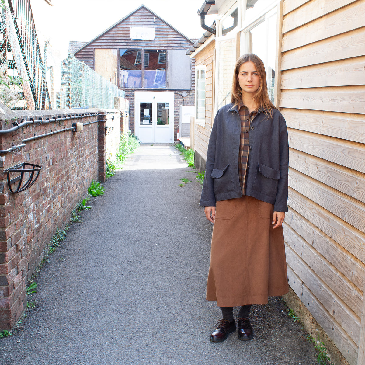 A woman stands in an outdoor alleyway wearing a dark charcoal utility jacket over a brown and black plaid shirt, paired with a brown skirt and black shoes.