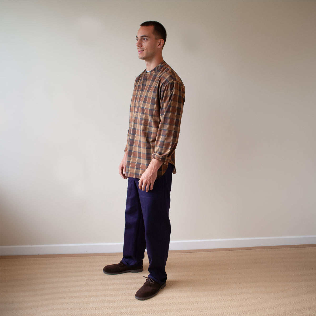 A man wearing a brown and orange plaid long-sleeve shirt, navy blue pants, and brown shoes, standing in profile against a plain beige wall and light wood floor.