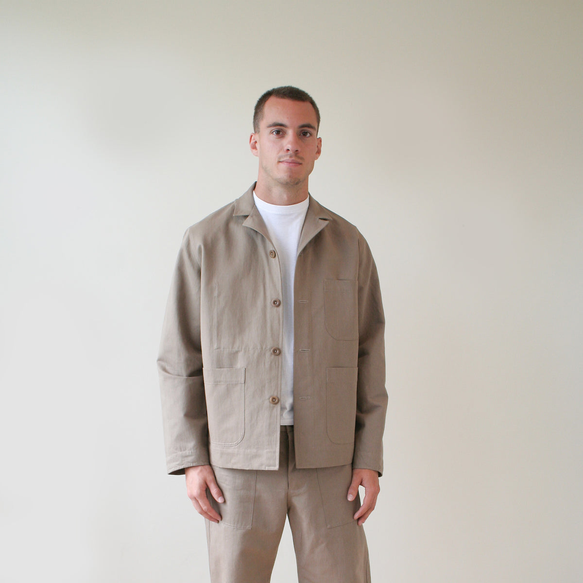 A man wearing a light beige utility jacket over a white t-shirt and matching beige trousers, standing against a plain off-white background.