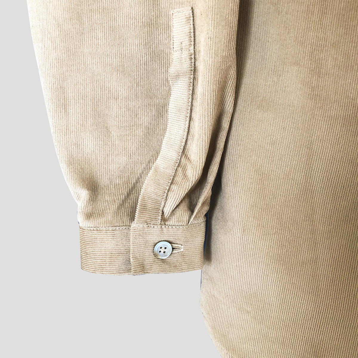 Close-up of a beige corduroy garment sleeve with a buttoned cuff, shown against a light gray background.