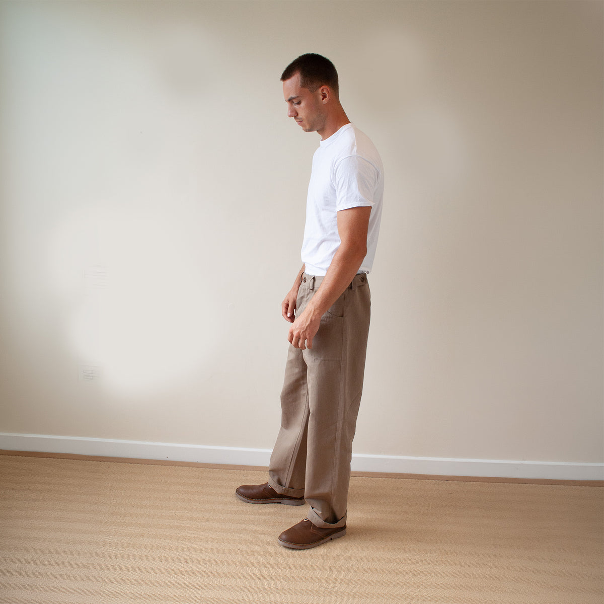 A man standing in profile wearing a white t-shirt, beige wide-leg trousers, and brown slip-on shoes in a minimalist room with a beige wall and light wood floor.