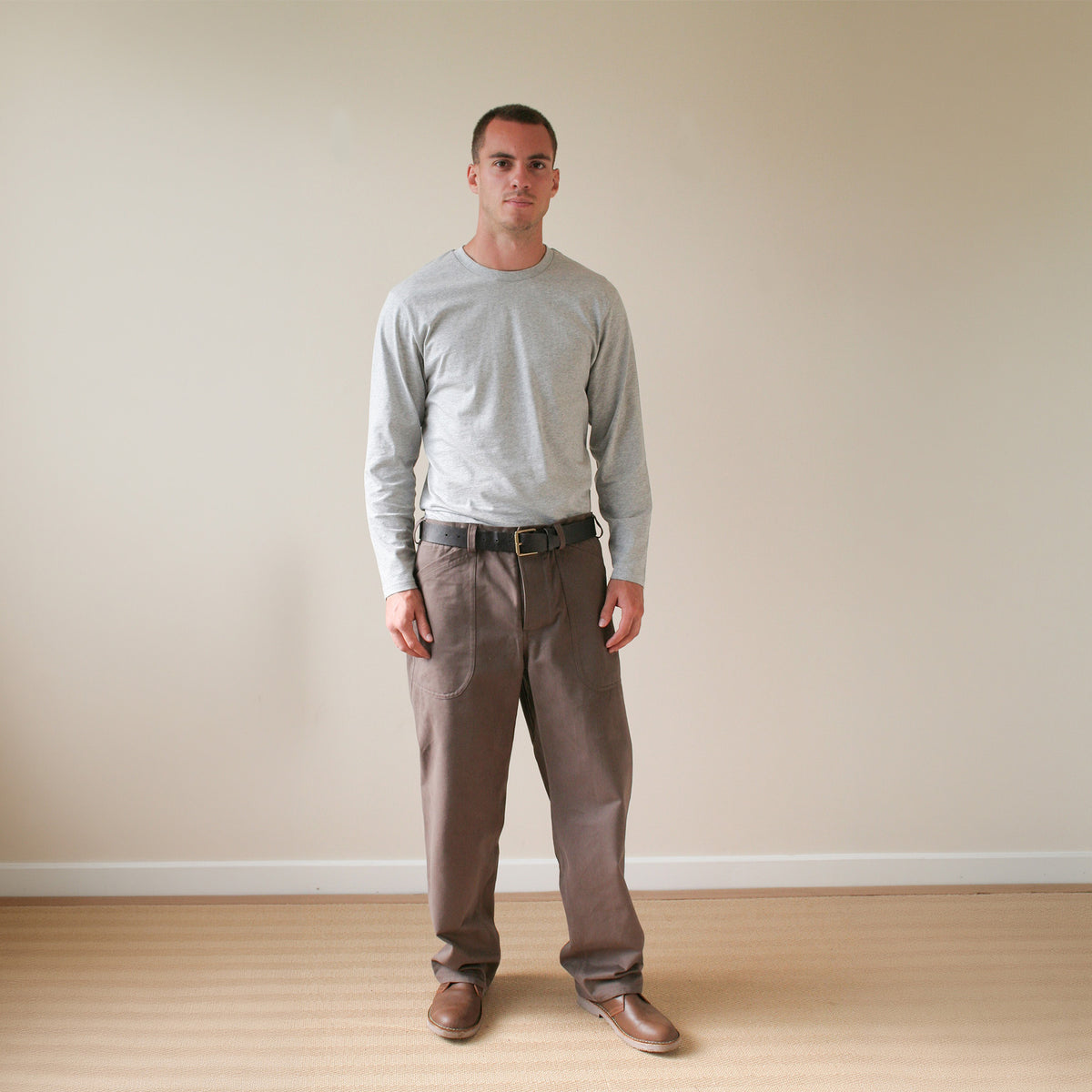 A man wearing a light gray long-sleeve shirt, brown trousers, and brown shoes stands against a plain beige wall and light wood floor.