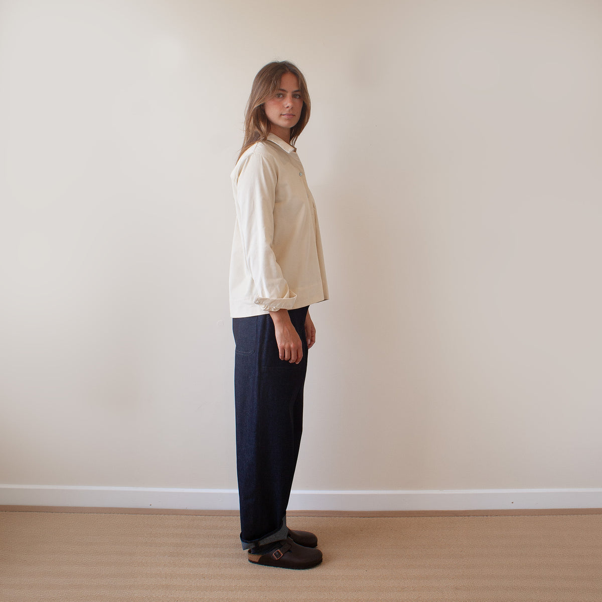A woman stands in profile wearing a cream long-sleeve button-up shirt, dark navy wide-leg trousers, and dark brown slip-on shoes against a plain beige wall and light wood floor.