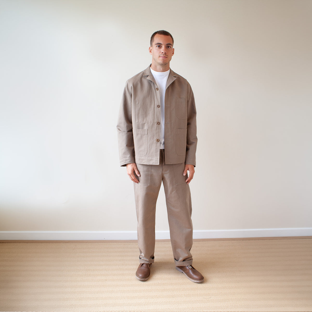 A man stands in a minimalist room with a beige wall and light wood floor, wearing a matching beige linen jacket and trousers set over a white t-shirt and brown shoes.