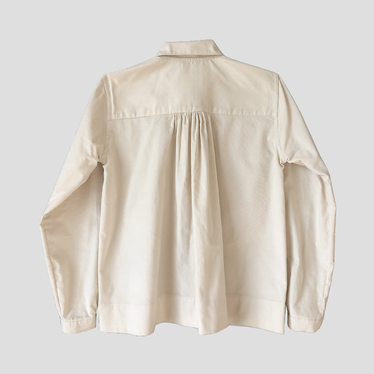 An off-white long-sleeve button-up shirt with a pleated back detail, shown from the back against a solid light gray background.
