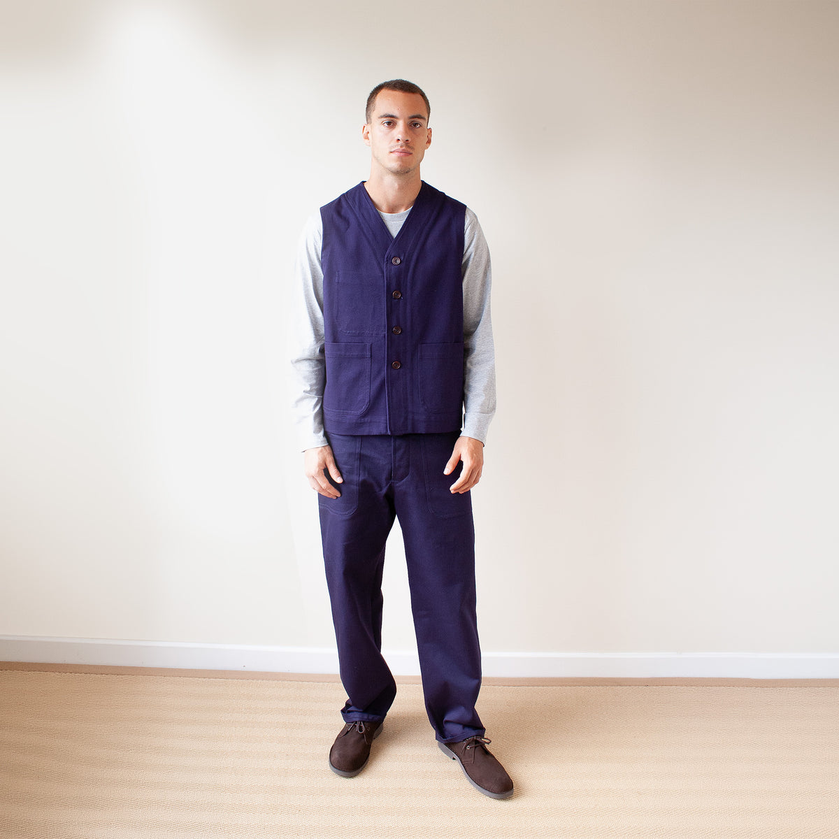 A man wearing a navy blue vest and matching trousers over a light gray long-sleeve shirt, standing in a minimalist room with a beige wall and light wood floor.