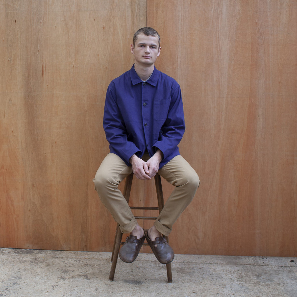 a young man standing against a wooden wall wearing a traditional chore jacket made in a sustainable tan brushed cotton canvas chore jacket in indigo worn by a young man sitting on a stool