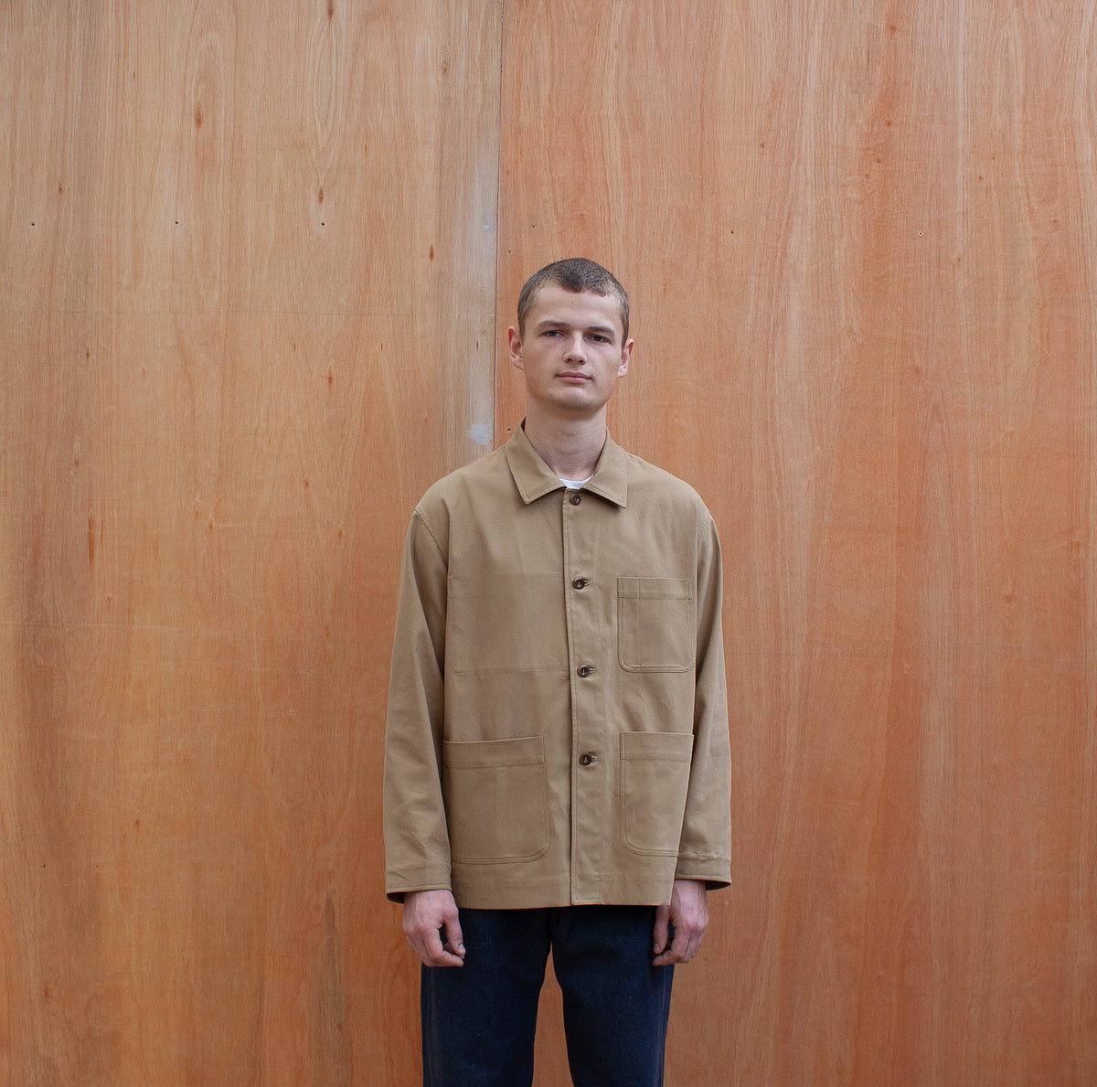 a young man standing against a wooden wall wearing a traditional chore jacket made in a sustainable tan brushed cotton canvas