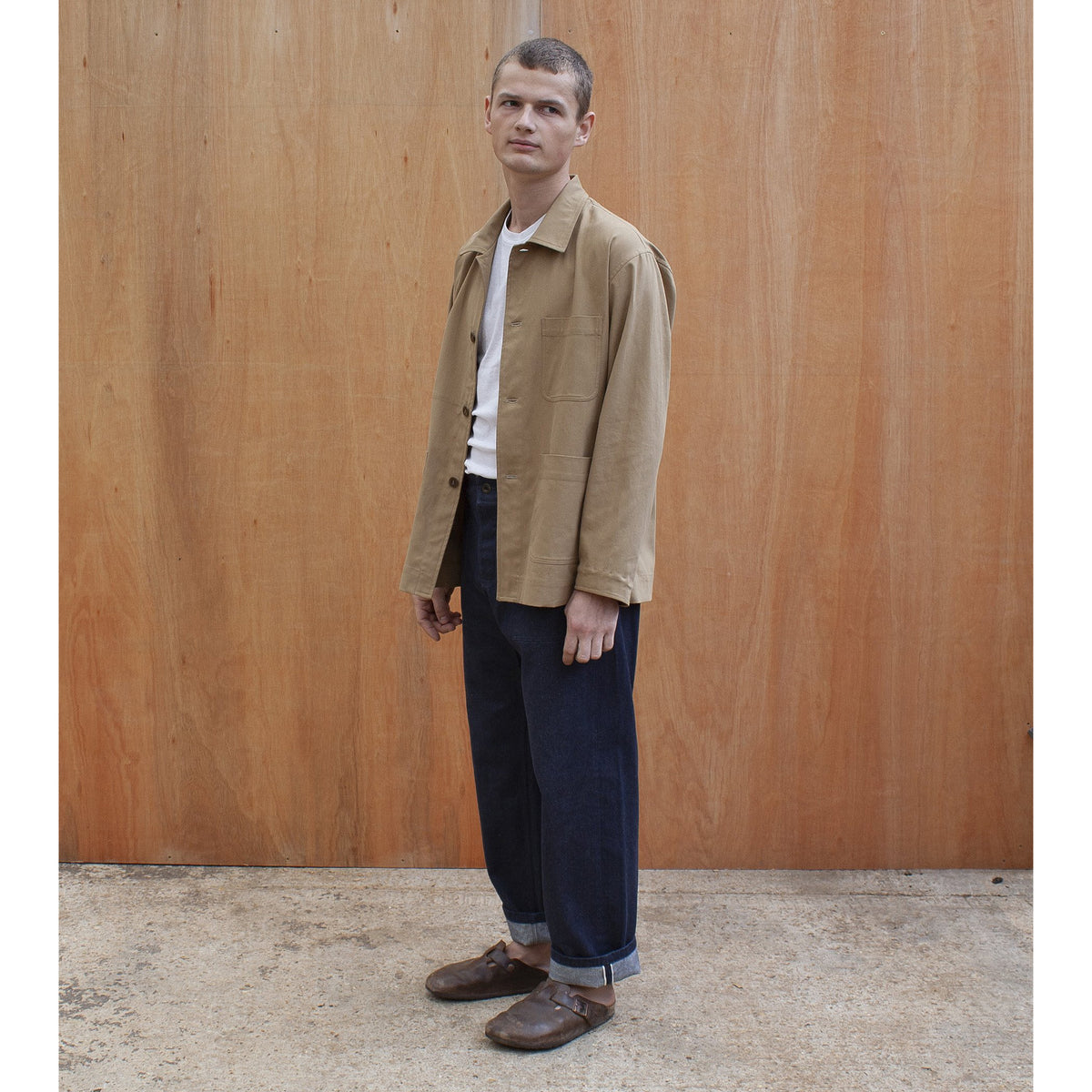 a young man standing against a wooden wall wearing a traditional chore jacket made in a sustainable tan brushed cotton canvas Edit alt text