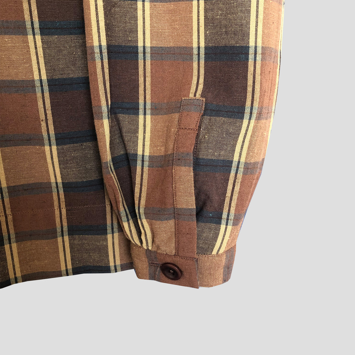 Close-up of a plaid shirt sleeve showing a button cuff and pocket detail, with a brown, beige, and navy pattern on a light gray background.