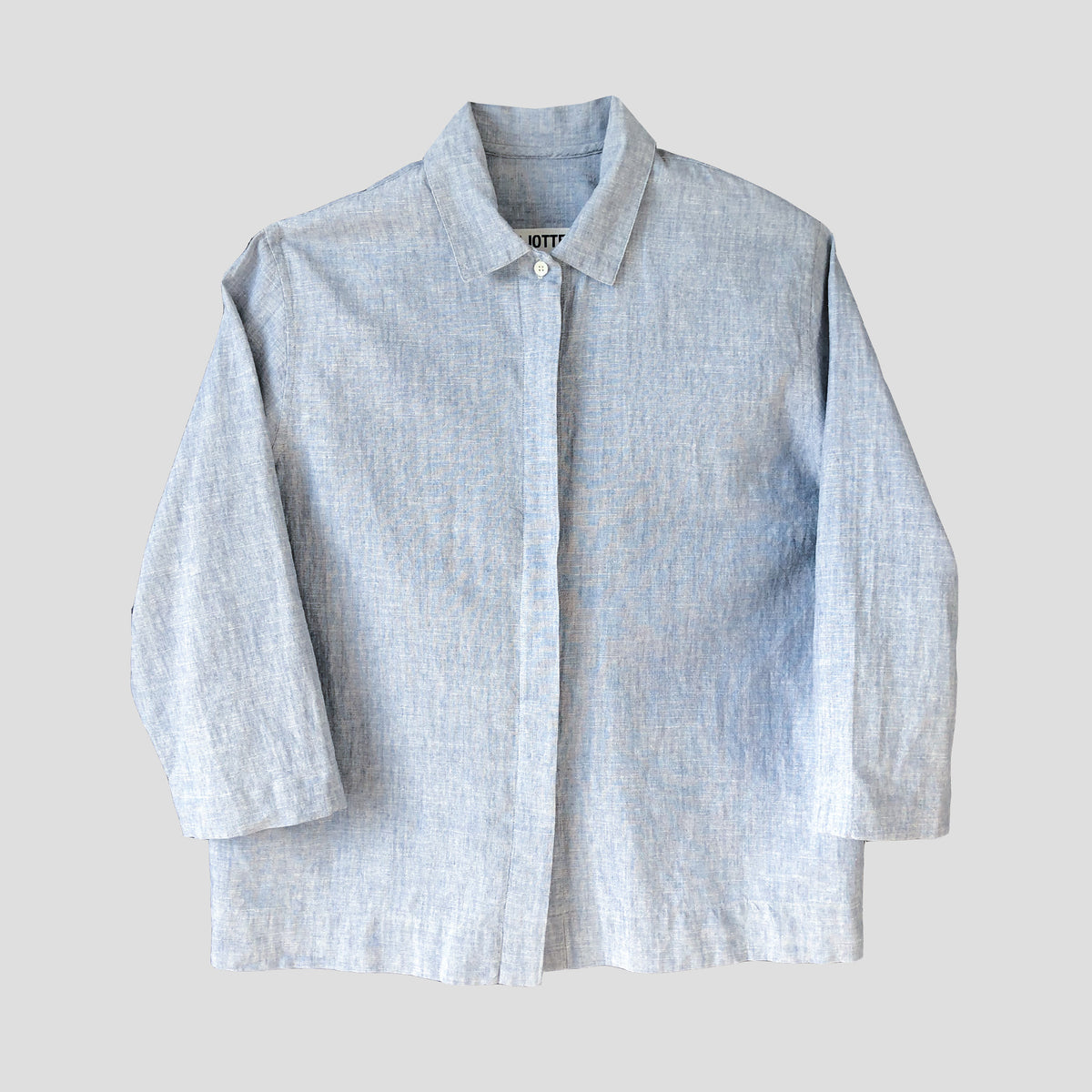 A light blue textured linen button-up shirt with a pointed collar and three-quarter sleeves, laid flat against a neutral light gray background.