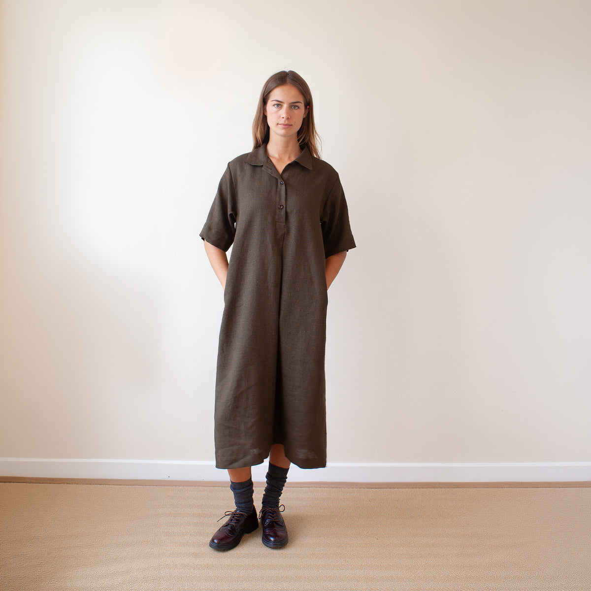 A woman stands in a minimalist room wearing a dark green short-sleeve linen shirt dress with a button-down front and side pockets.