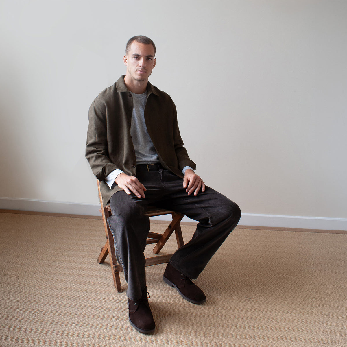 A man sits on a wooden folding chair in a minimalist room, wearing a dark olive green jacket over a gray t-shirt, dark charcoal trousers, and dark brown leather shoes.