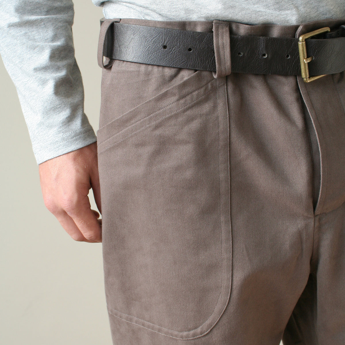 Close-up of a person wearing taupe-brown canvas utility pants with a black leather belt and gold buckle, paired with a light gray long-sleeve shirt.