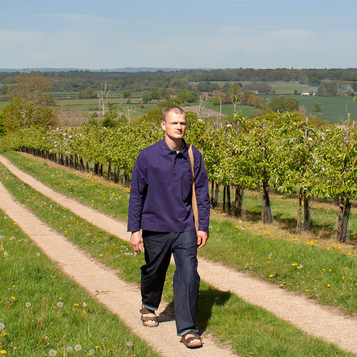 Man walking along a dirt path in a vineyard with greenery and trees in the background.