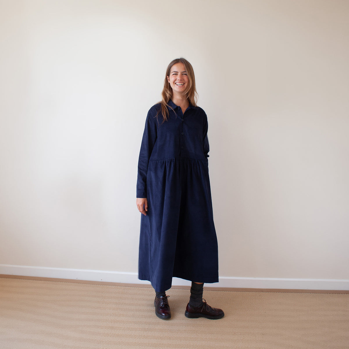A woman stands in a minimalist room with a beige wall and light wood floor, wearing a long-sleeved navy blue midi dress with a gathered waist, dark socks, and dark brown shoes.