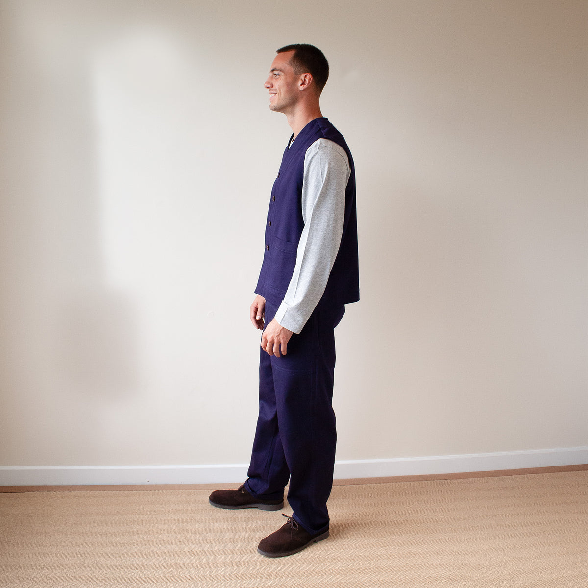 A man stands in profile wearing a navy blue waistcoat and matching trousers over a light gray long-sleeve shirt, paired with dark brown shoes, against a plain beige wall and light wood floor.