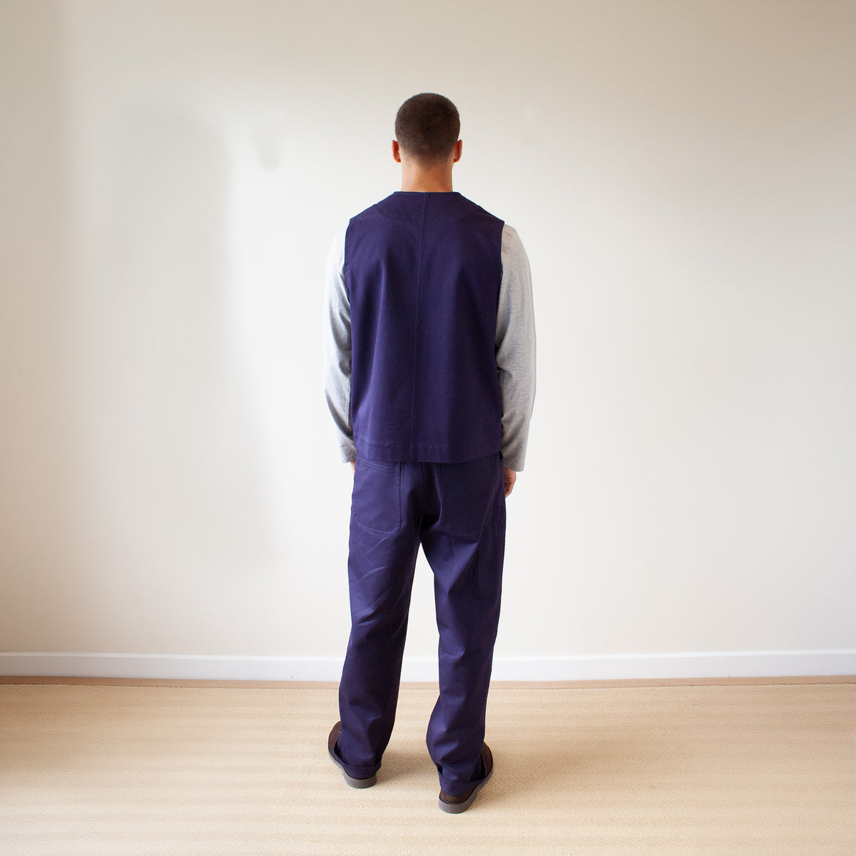 A man viewed from behind wearing a navy blue scrub-style top and matching pants over a light gray long-sleeve shirt, standing on a light wood floor against a plain off-white wall.