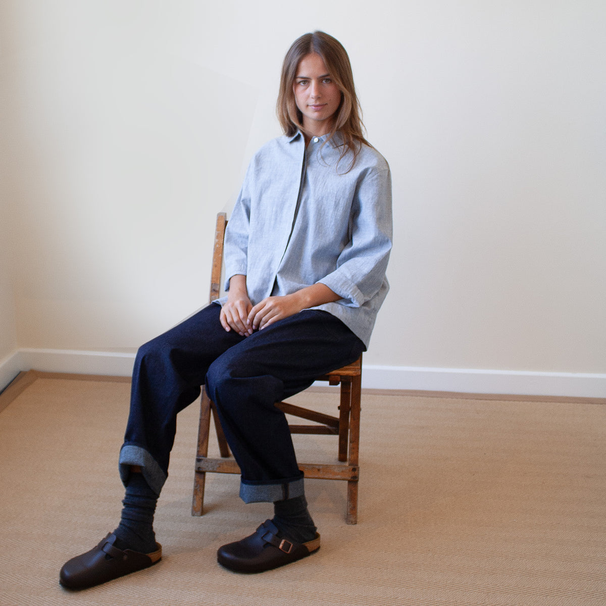 A woman sits on a wooden chair in a minimalist room, wearing a light blue button-up shirt, dark navy cuffed jeans, dark socks, and dark brown clogs.