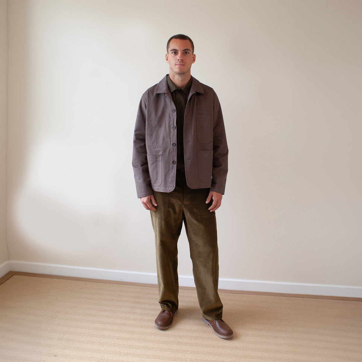 A man stands in a minimalist room with beige walls and light wood flooring, wearing a muted brown utility jacket over a dark shirt, olive green corduroy pants, and brown leather shoes.