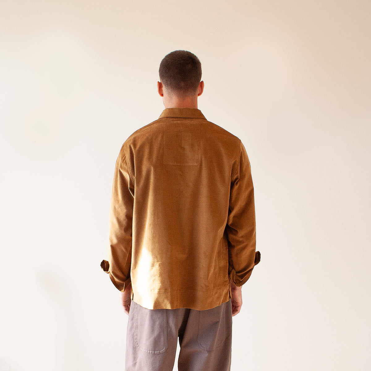 A man viewed from behind wearing a brown corduroy button-down shirt and gray trousers, standing against a plain off-white background.