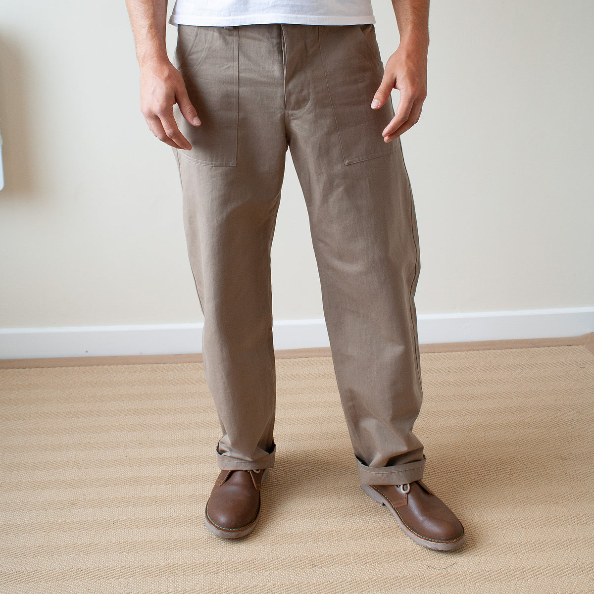 A person wearing straight-leg brown trousers and brown leather shoes, standing indoors against a light beige wall and textured floor.