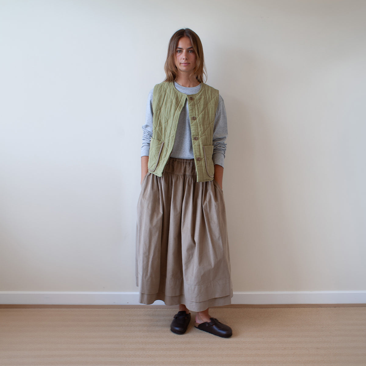 A woman stands in a minimalist room with a beige wall and light wood floor, wearing a sage green quilted vest over a light gray long-sleeve top and a beige wide-leg skirt, paired with dark brown loafers.