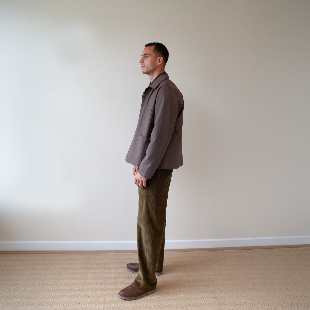 A man standing in profile wearing a taupe-brown jacket, olive green corduroy trousers, and brown shoes, set against a plain beige wall and light wood floor.