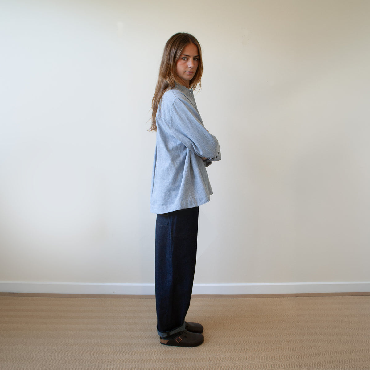 A woman stands in profile wearing an oversized light blue long-sleeve shirt and dark navy wide-leg jeans, standing on a light wood floor against a plain off-white wall.