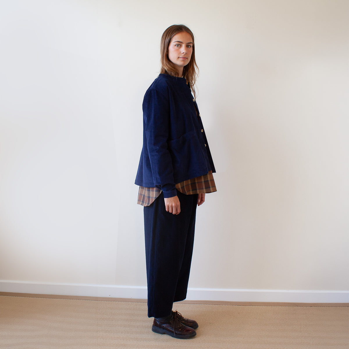A woman stands in profile wearing a dark navy corduroy jacket over a brown and beige plaid shirt, paired with dark navy wide-leg trousers and dark brown leather shoes, against a plain off-white wall and light wood floor.