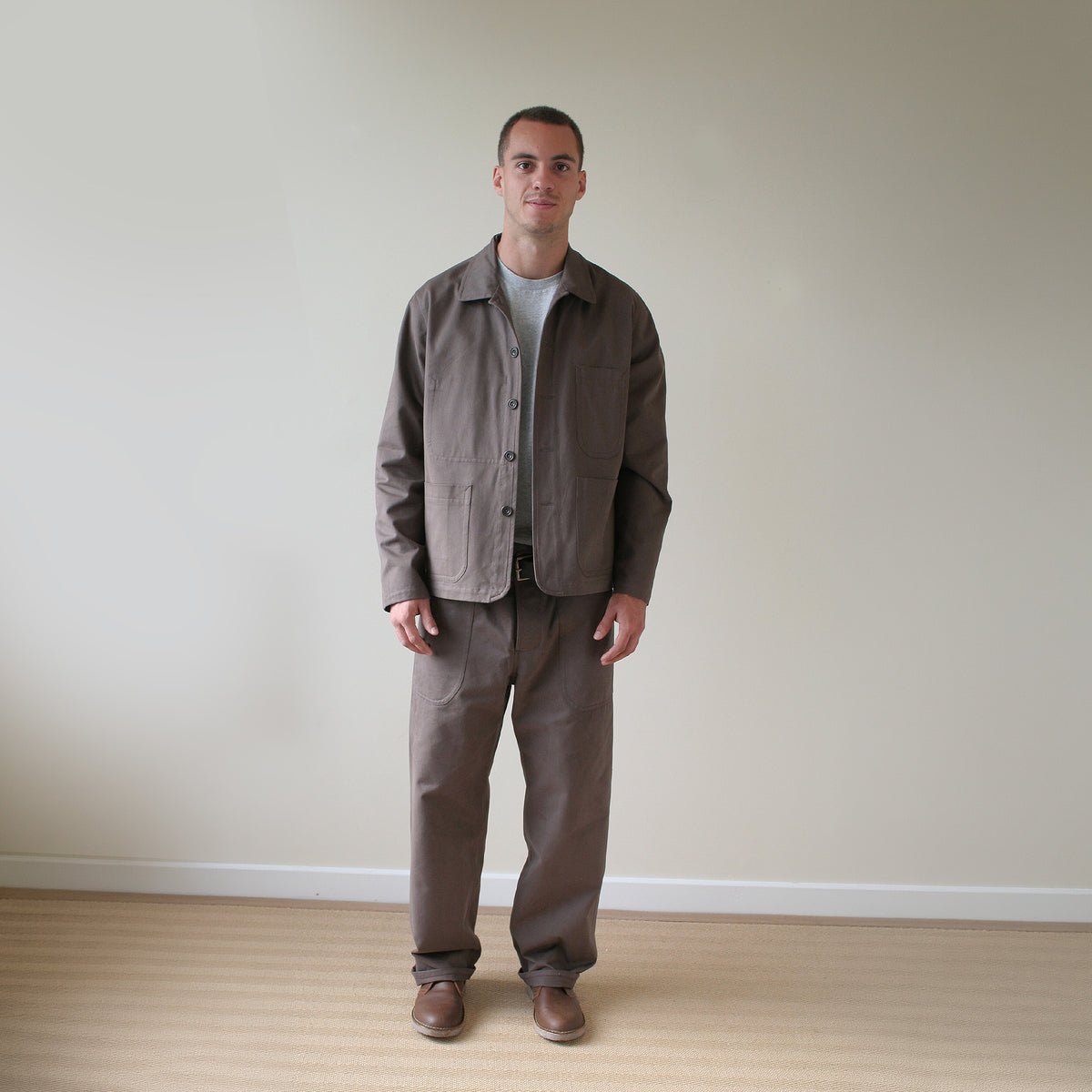 A man stands in a minimalist room with a beige wall and light wood floor, wearing a brown utility jacket and matching brown pants over a light gray t-shirt.