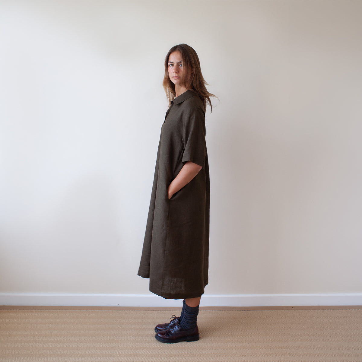 A woman stands in profile wearing a loose, oversized short-sleeve shirt dress in dark brown, paired with dark socks and dark brown leather shoes, set against a plain off-white wall and light wood floor.