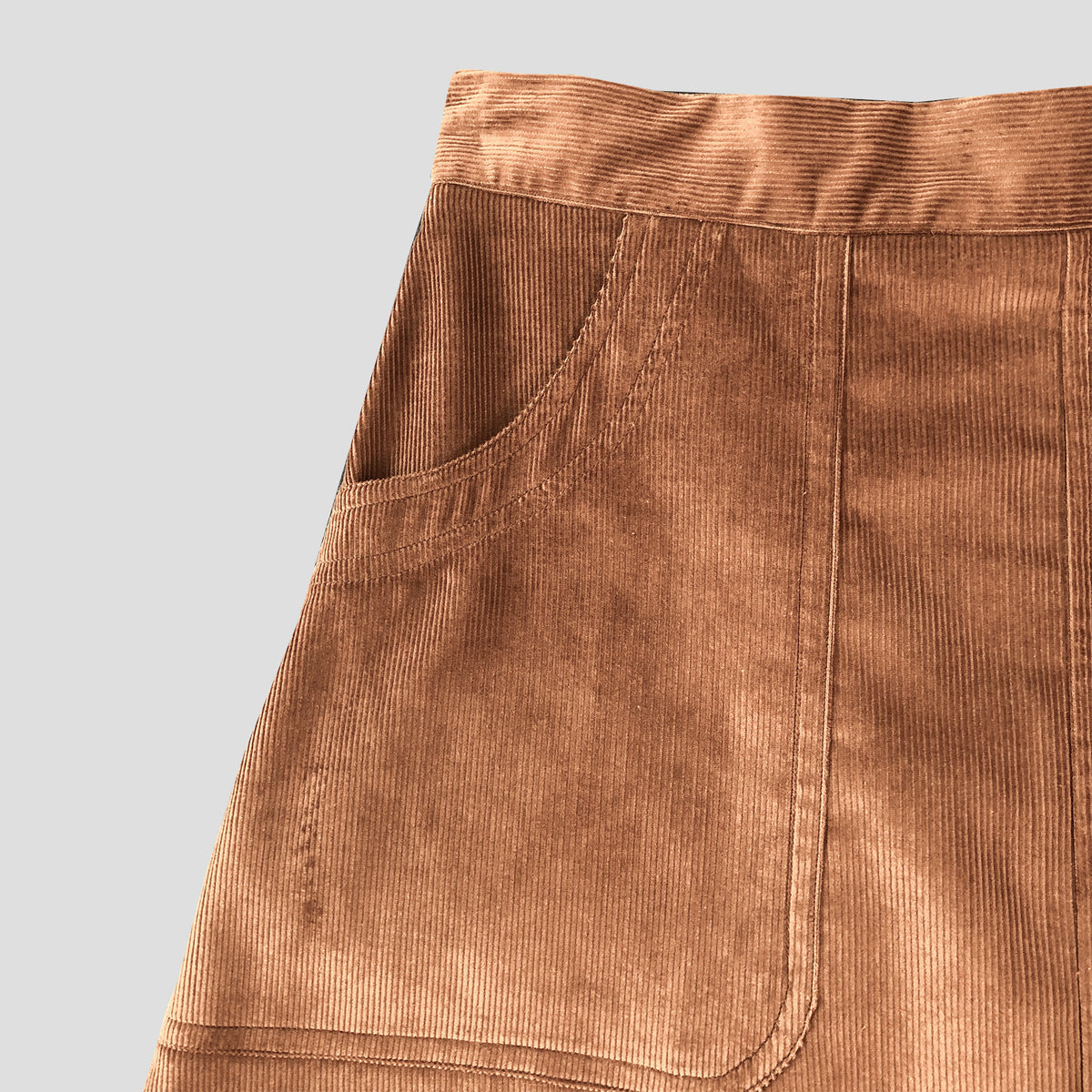 Close-up of a brown corduroy garment, showing a side pocket and waistband against a light gray background.