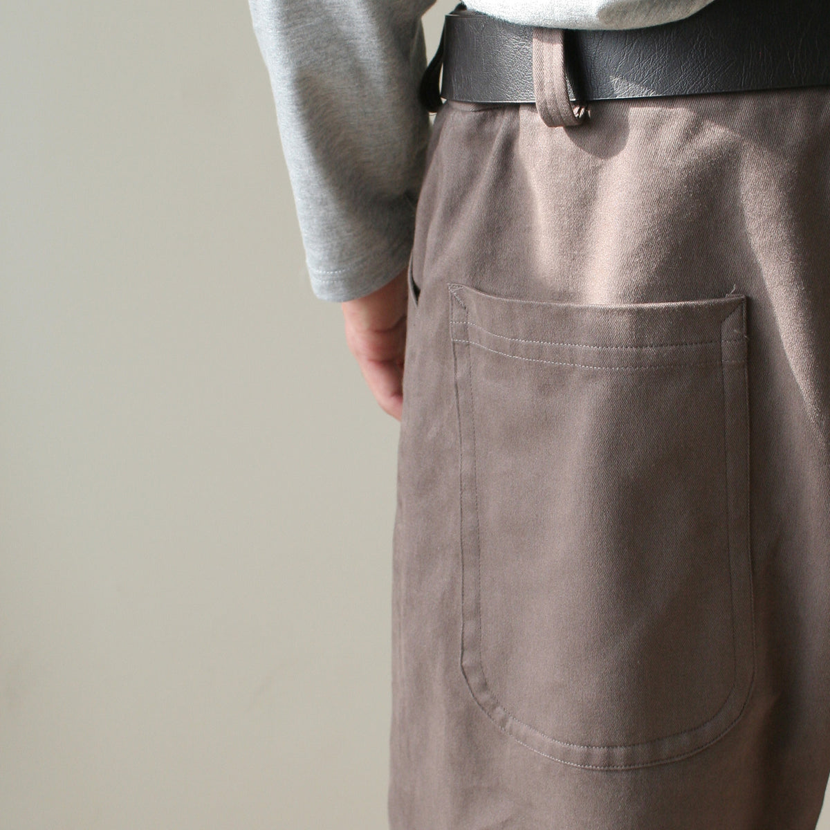 Close-up of a person wearing taupe-brown corduroy trousers with a large back pocket, a black belt, and a light gray long-sleeve shirt against a plain beige background.